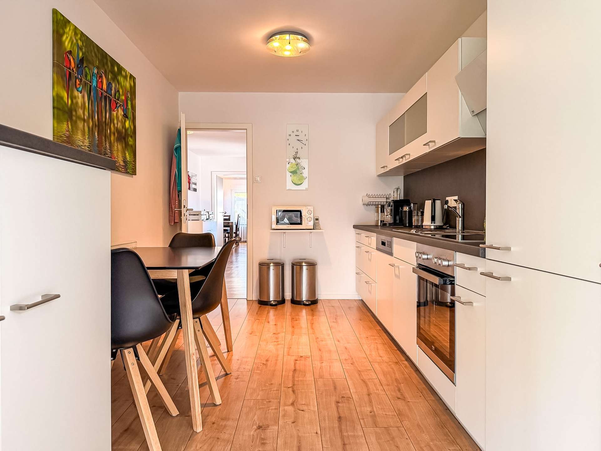 Bright modern galley kitchen with white cabinets, stainless steel appliances, a small dining table and black chairs at the left.
