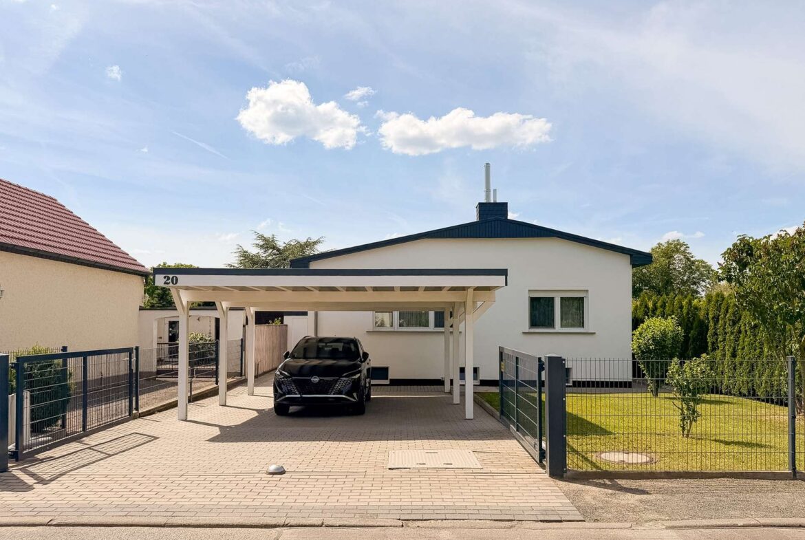White single-story house with a blue gabled roof and a covered carport, a black SUV parked in the driveway, and a metal fence surrounding the property with a lawn to the right. The carport is labeled with the number 20 at the edge of the structure.