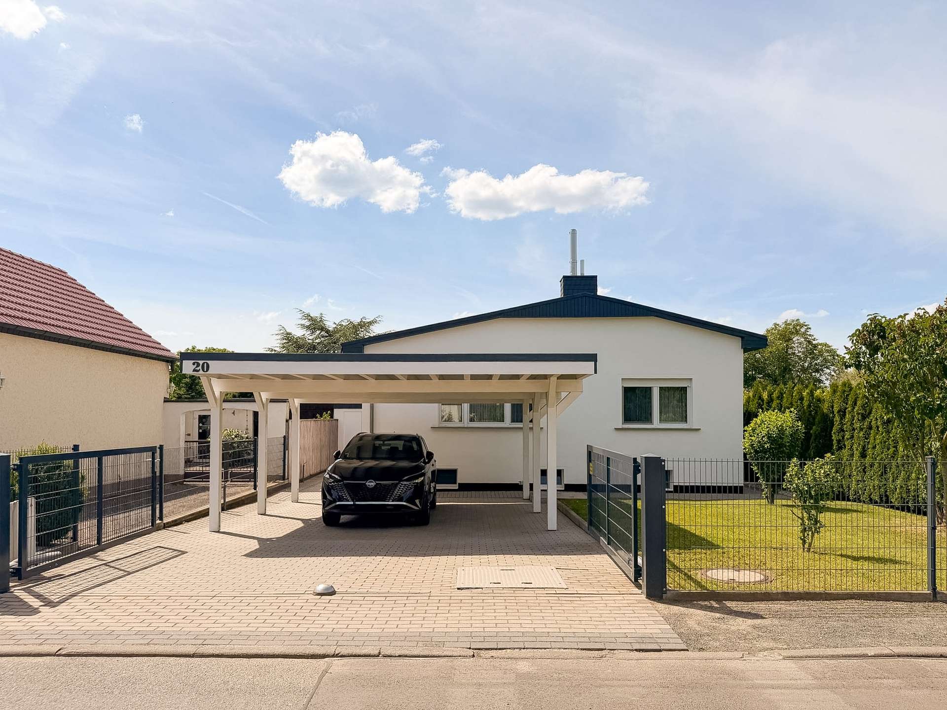 White single-story house with a blue gabled roof and a covered carport, a black SUV parked in the driveway, and a metal fence surrounding the property with a lawn to the right. The carport is labeled with the number 20 at the edge of the structure.