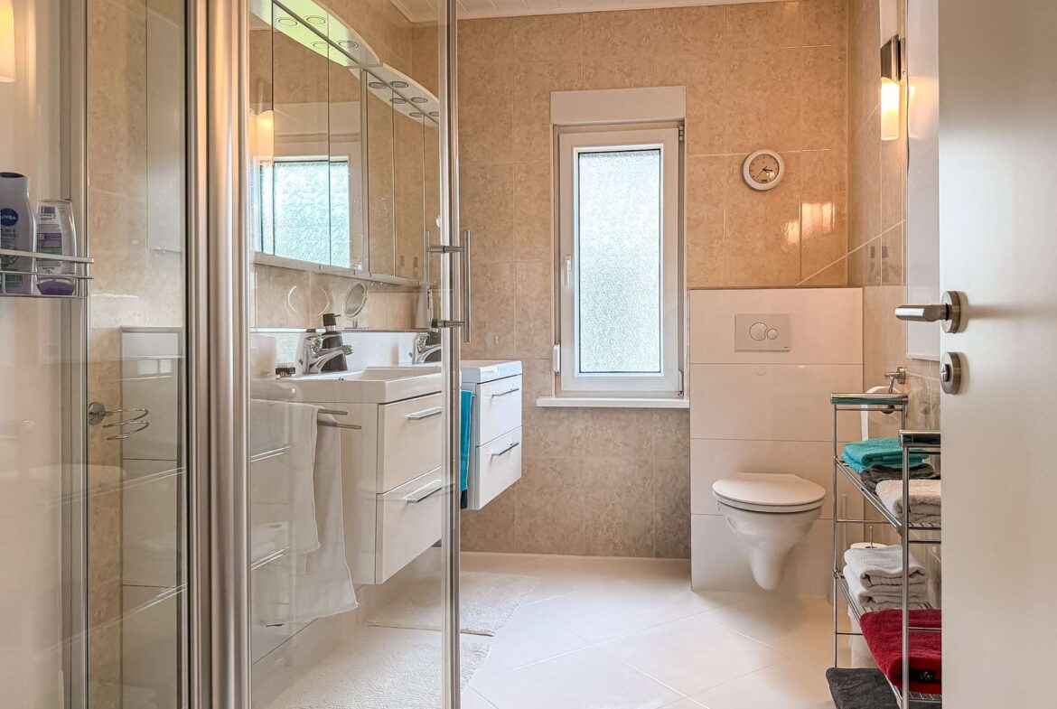 Modern bathroom with glass-walled shower on the left, white vanity with dual sinks, and a wall-mounted toilet; beige tiled walls and a small frosted window center-right.