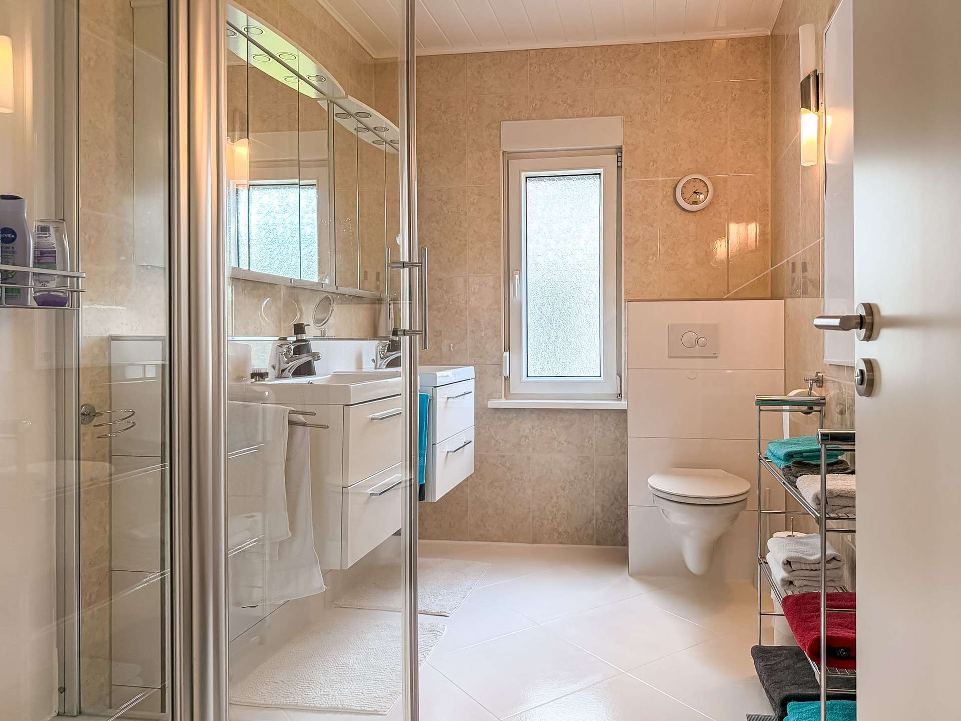 Modern bathroom with glass-walled shower on the left, white vanity with dual sinks, and a wall-mounted toilet; beige tiled walls and a small frosted window center-right.