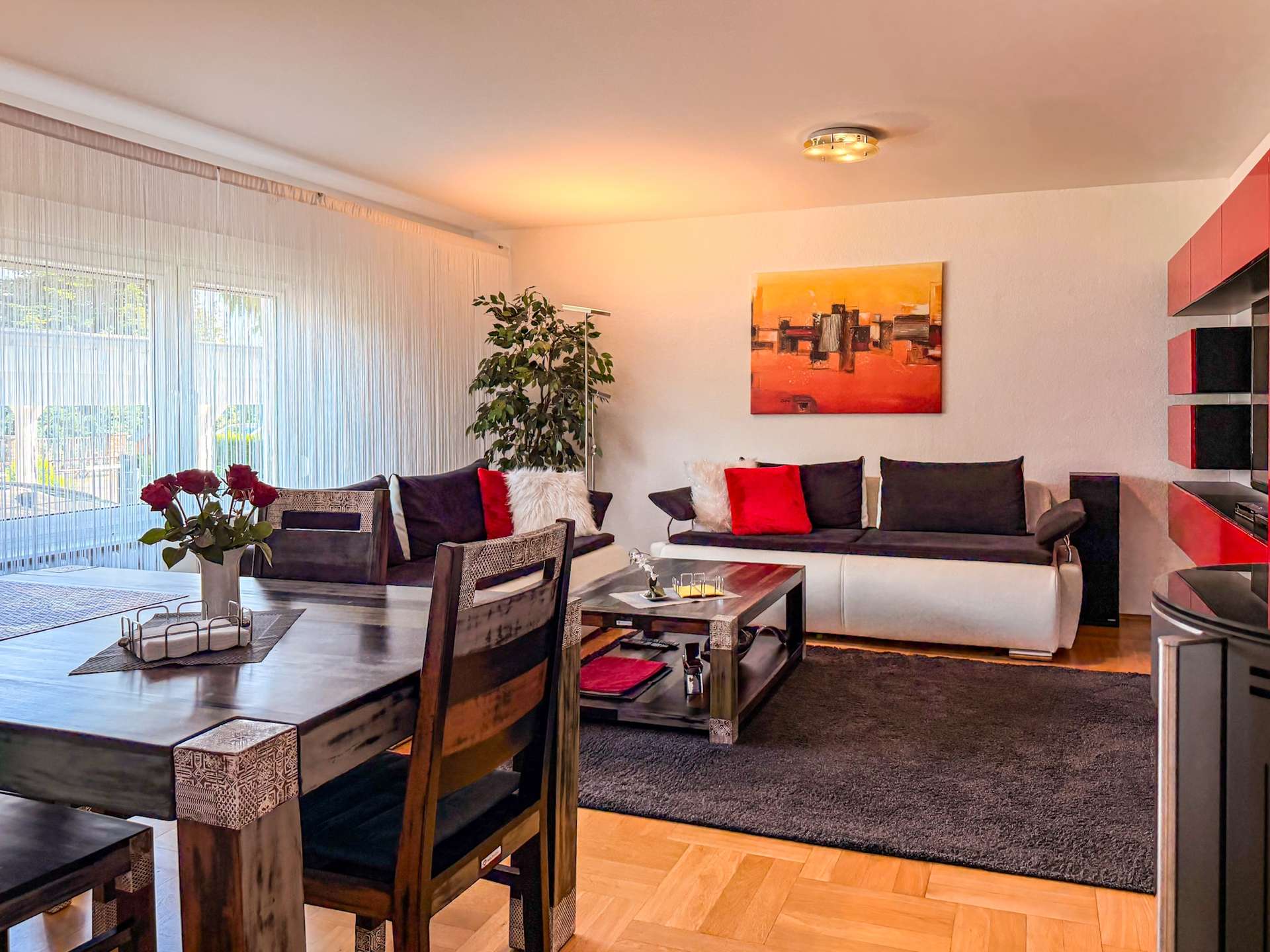 Bright living room with a dark wood dining set, red and black cushions, and an abstract orange painting on the wall.