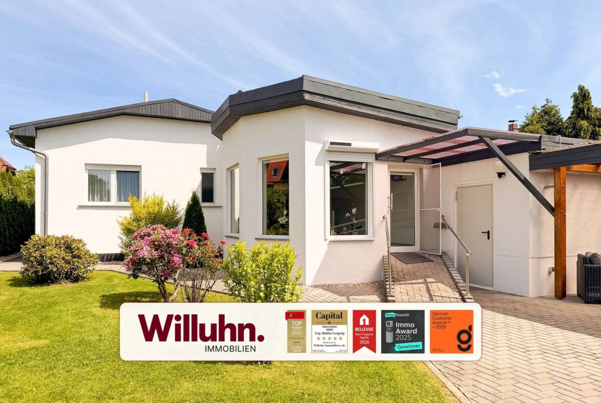 Front view of a white modern house with a glass entry and a large Willuhn Immobilien sign in the lawn, including award stickers.