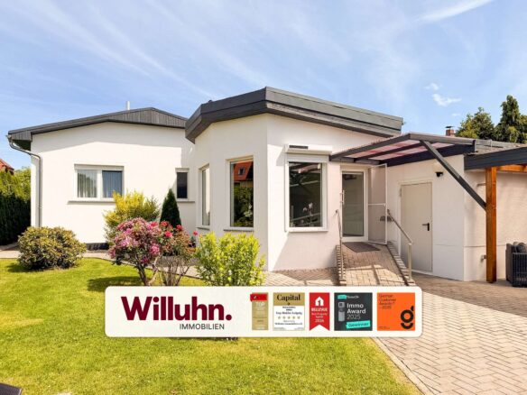 Front view of a white modern house with a glass entry and a large Willuhn Immobilien sign in the lawn, including award stickers.