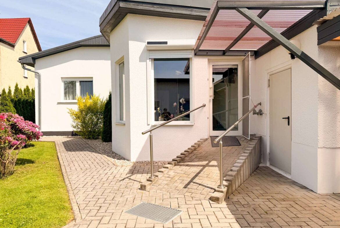 White modern house entrance featuring a glass door, brick ramp with stainless handrails, and a landscaped garden path on the left.
