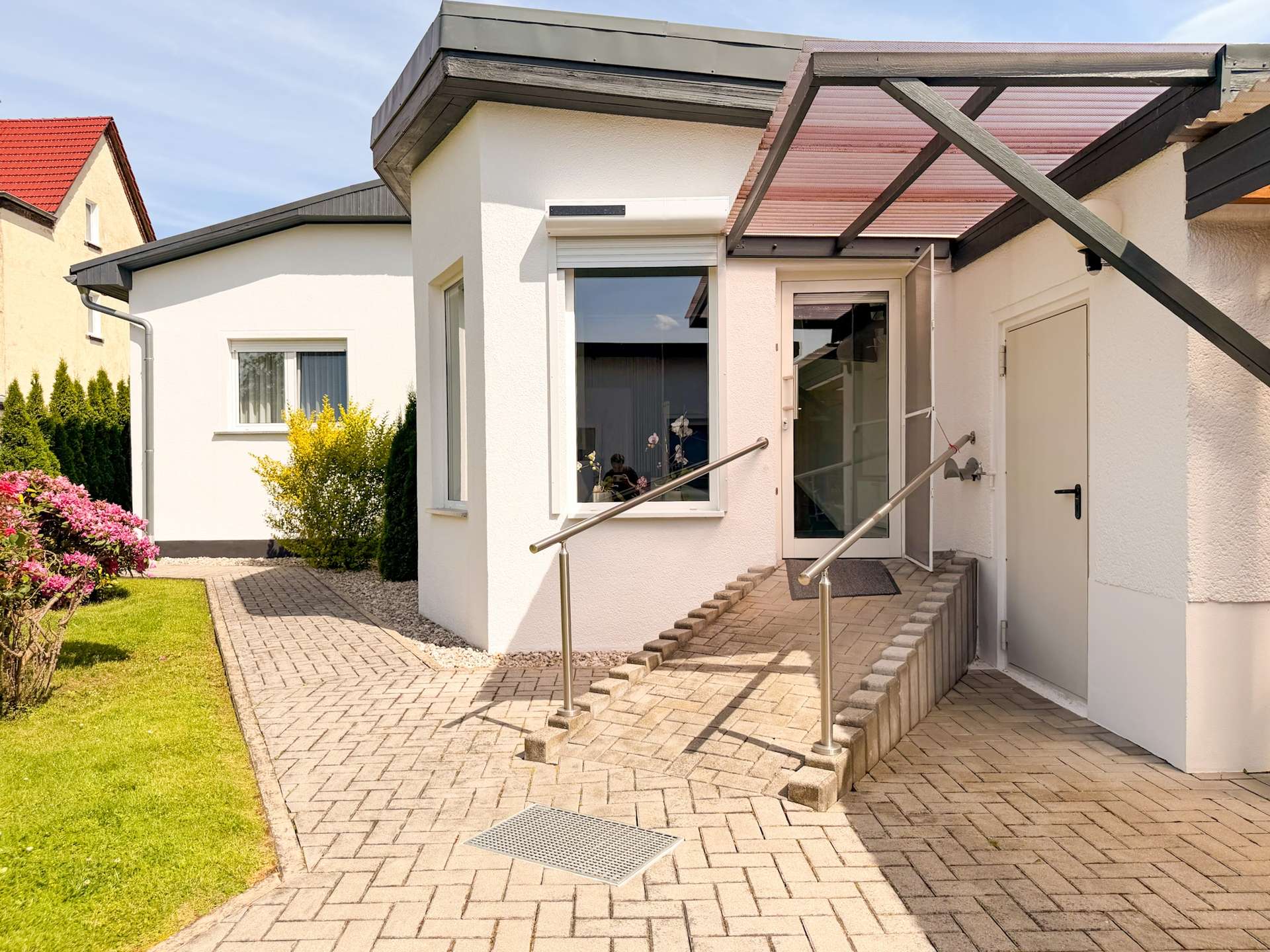 White modern house entrance featuring a glass door, brick ramp with stainless handrails, and a landscaped garden path on the left.