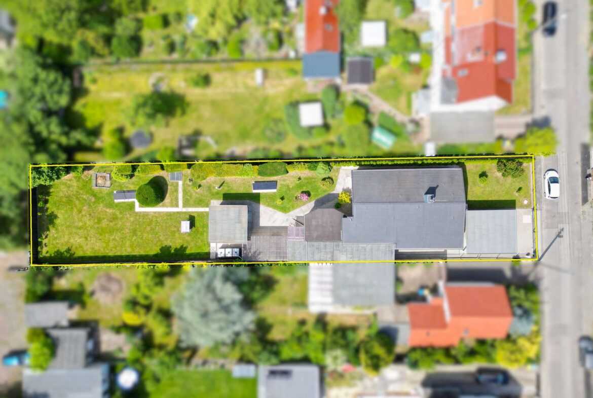 Aerial view of a long rectangular residential lot outlined in yellow, with a green lawn, garden features, and several buildings along a street.