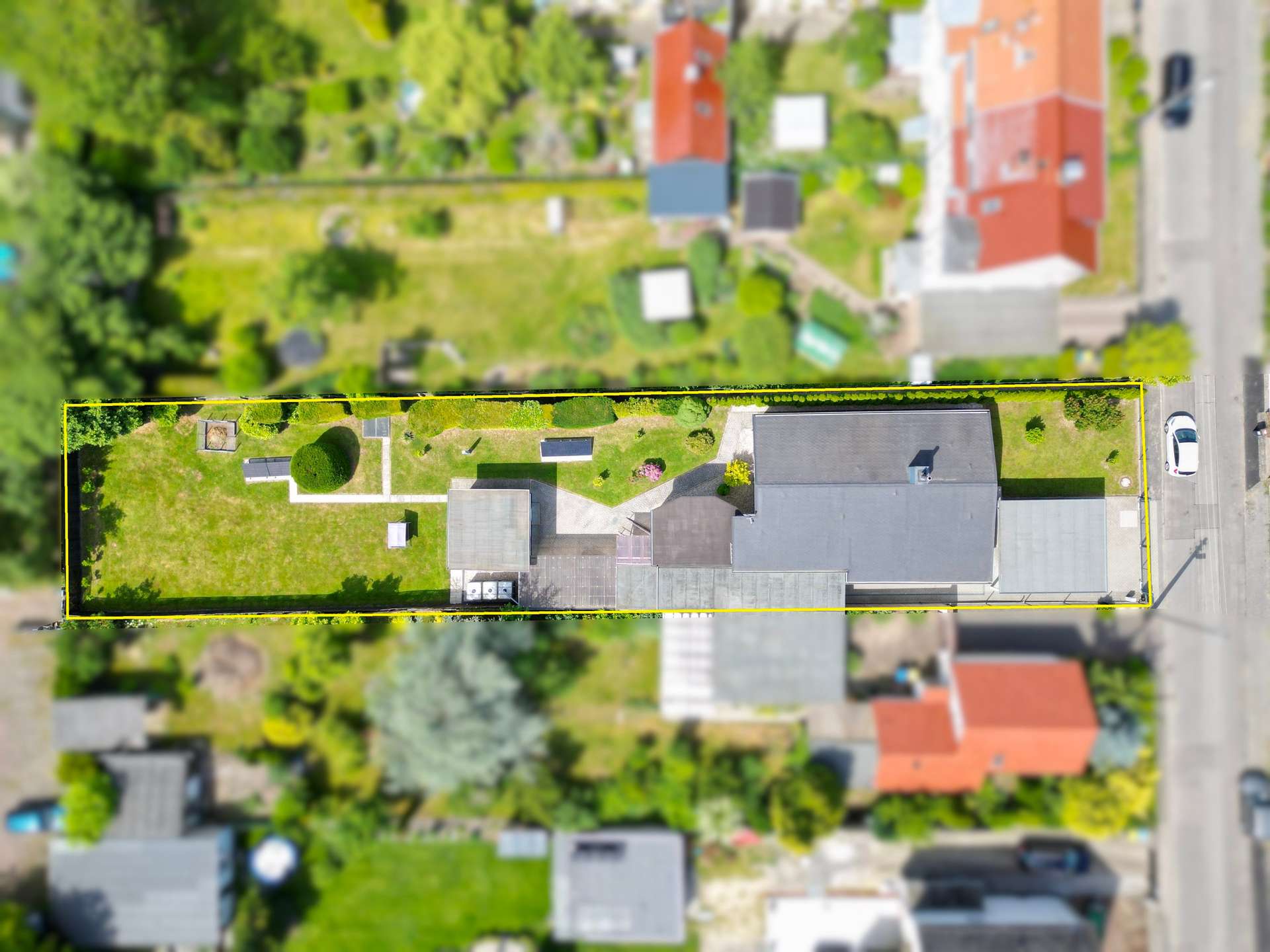 Aerial view of a long rectangular residential lot outlined in yellow, with a green lawn, garden features, and several buildings along a street.