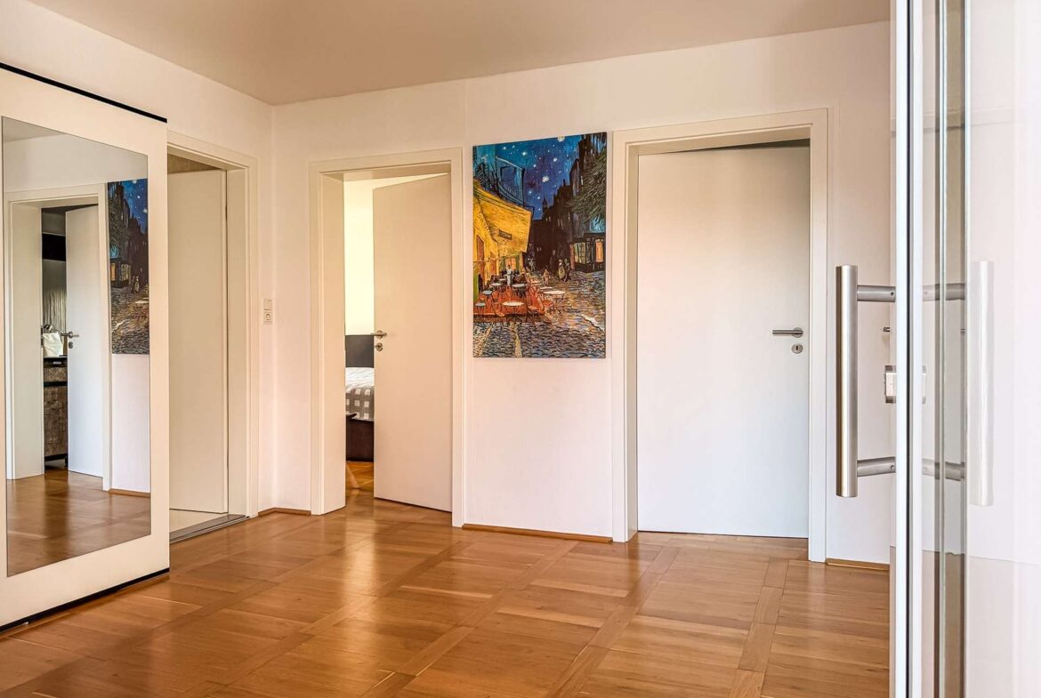 Bright hallway with white walls, a large framed painting of a night street scene, and a wooden parquet floor; glass doors on the right.