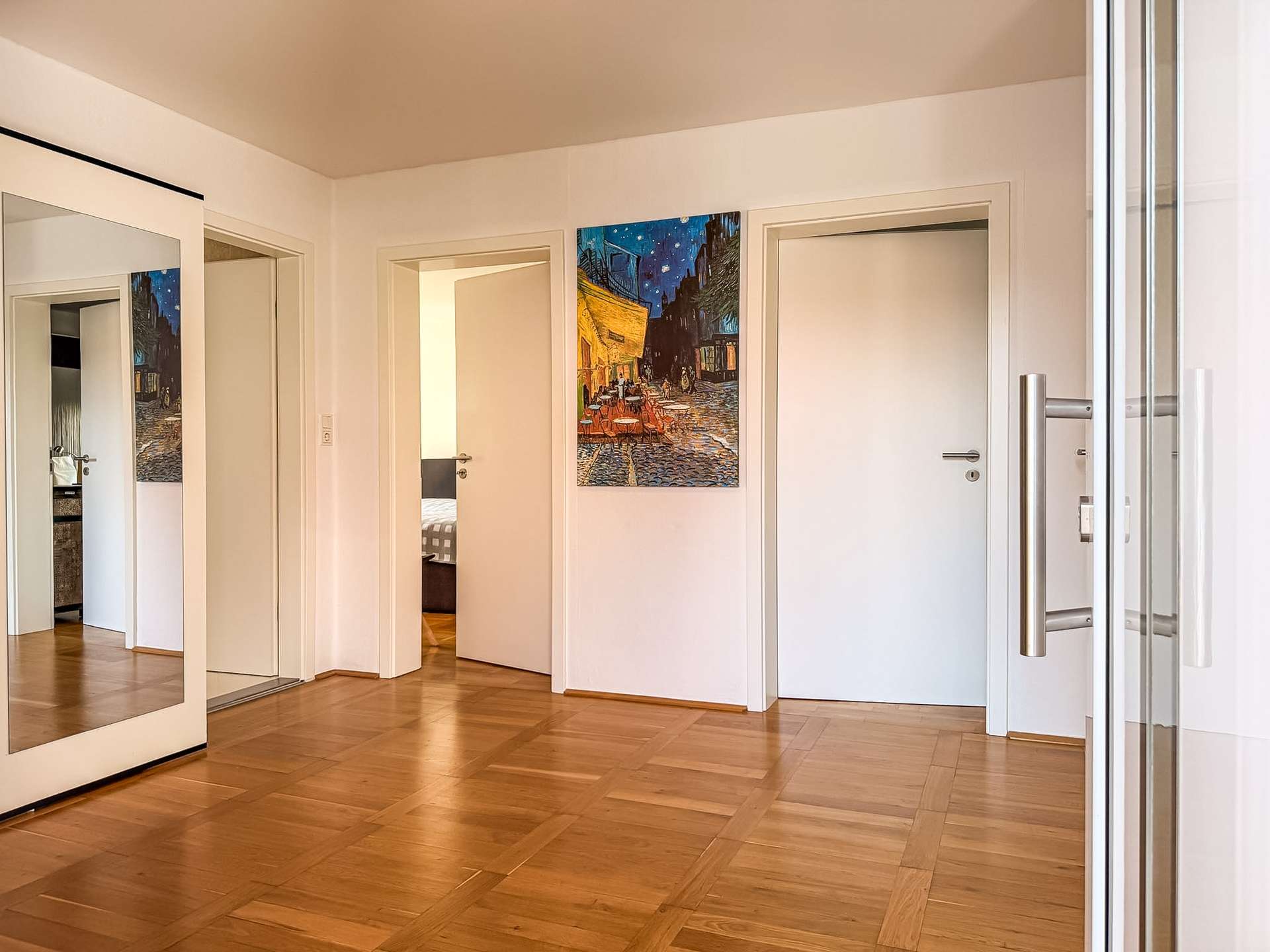 Bright hallway with white walls, a large framed painting of a night street scene, and a wooden parquet floor; glass doors on the right.