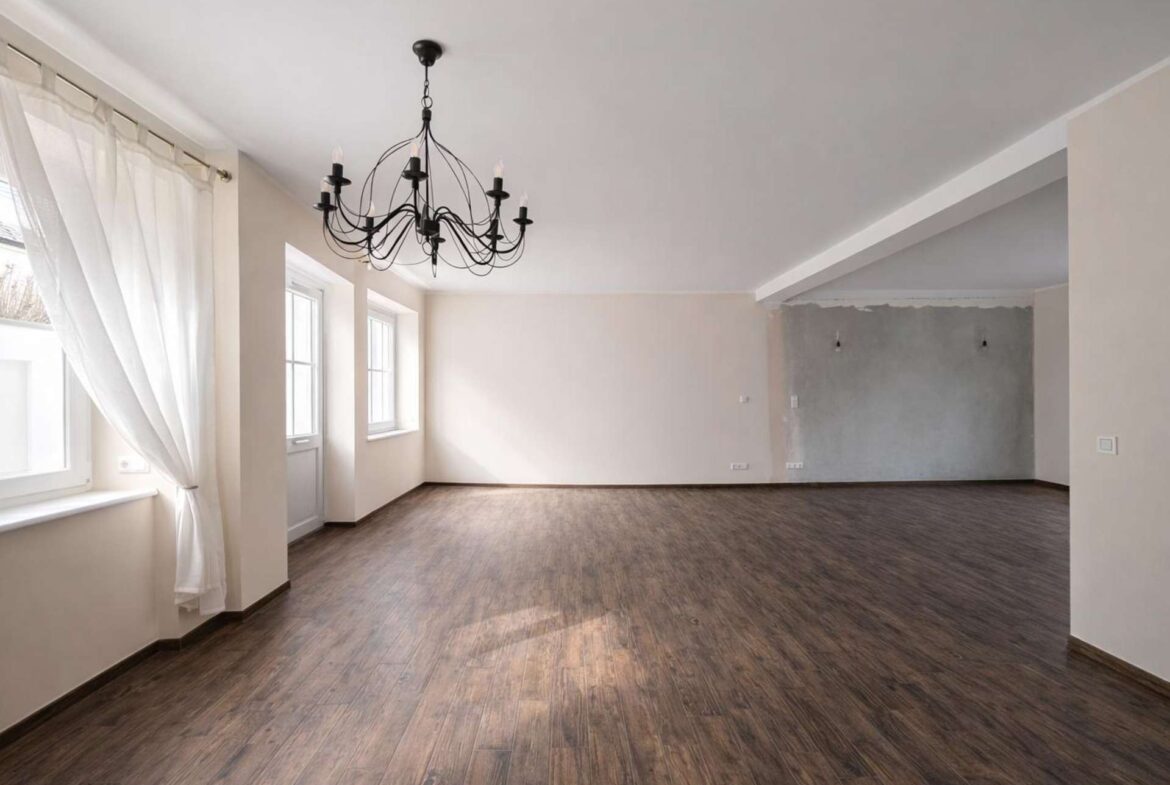 Empty living room with dark wood floors, a black chandelier, and large windows with sheer white curtains.