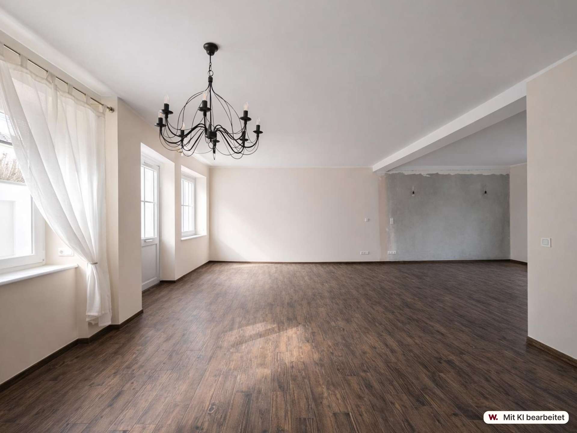Empty living room with dark wood floors, a black chandelier, and large windows with sheer white curtains.