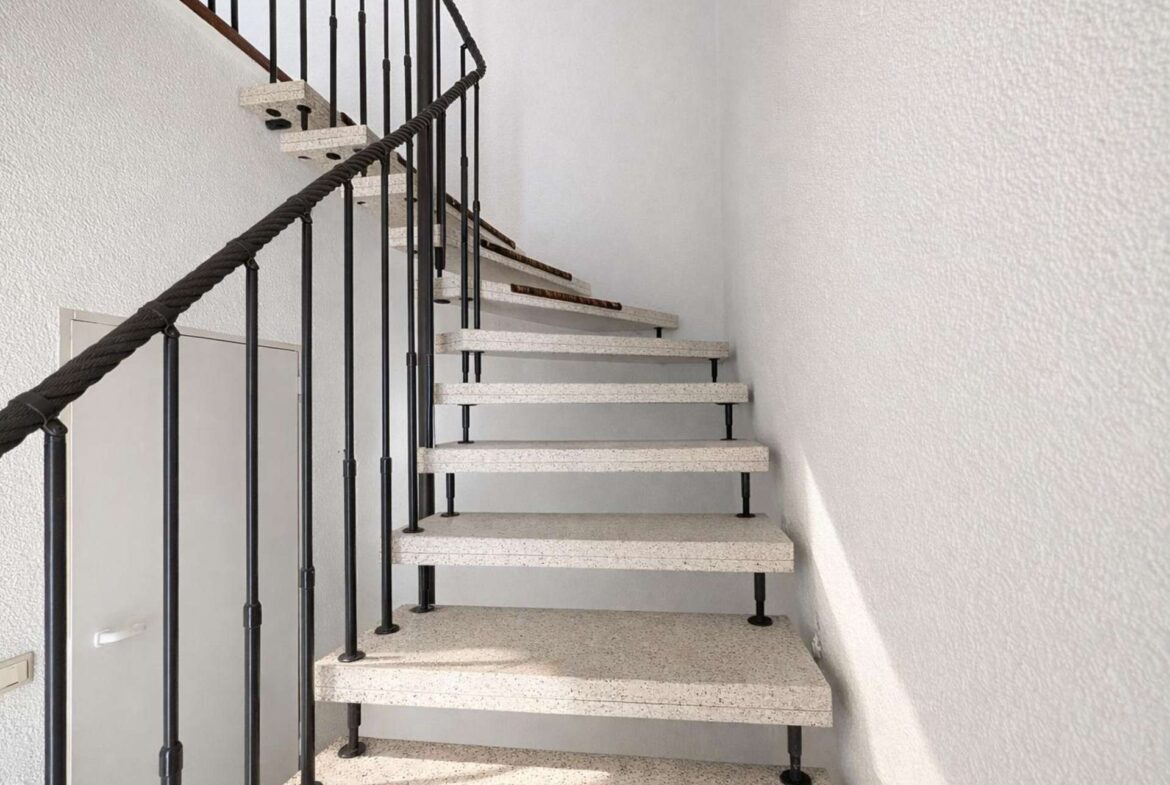 Interior spiral staircase with black metal railing and white speckled steps, ascending along white textured walls in a bright stairwell.