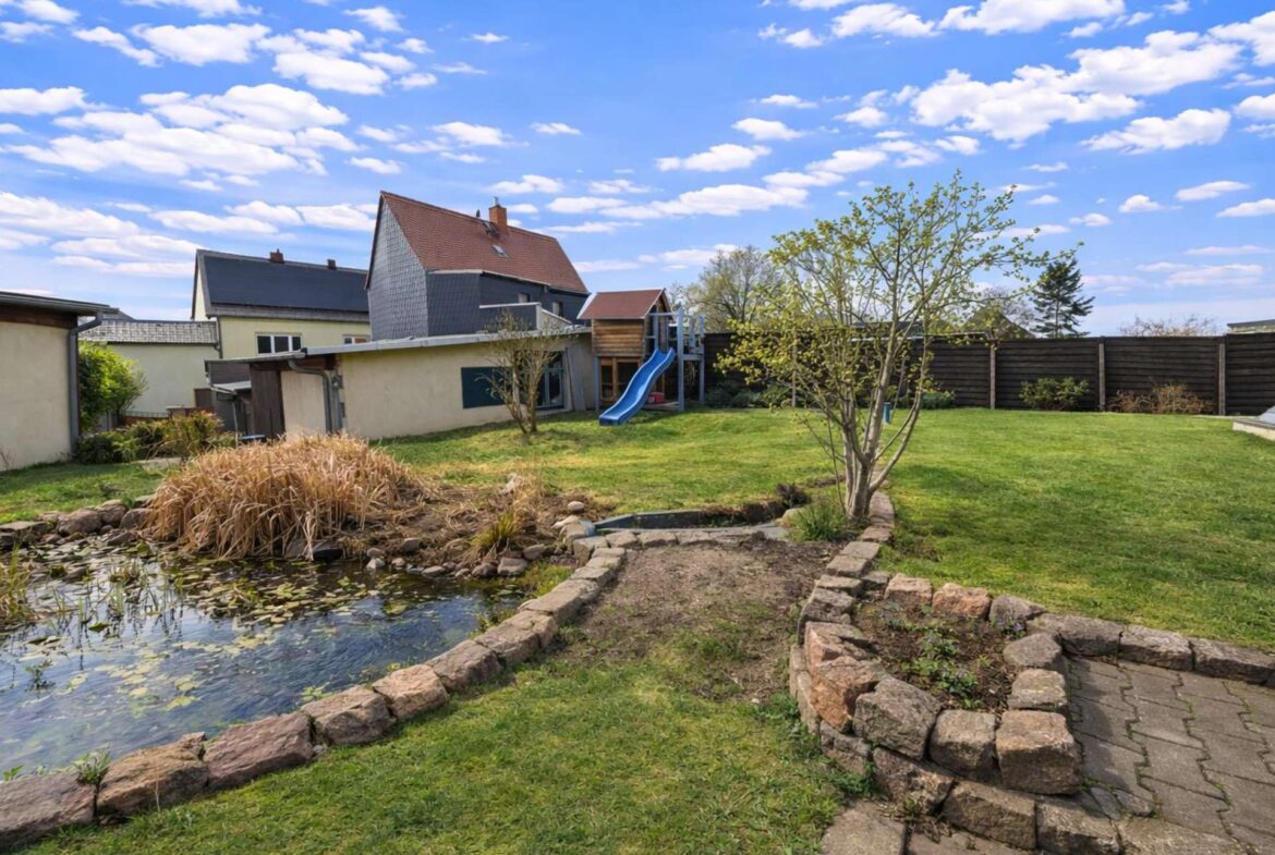 Backyard with a stone-edged pond, a blue playground slide, a small tree, and a wooden fence under a bright blue sky with clouds.