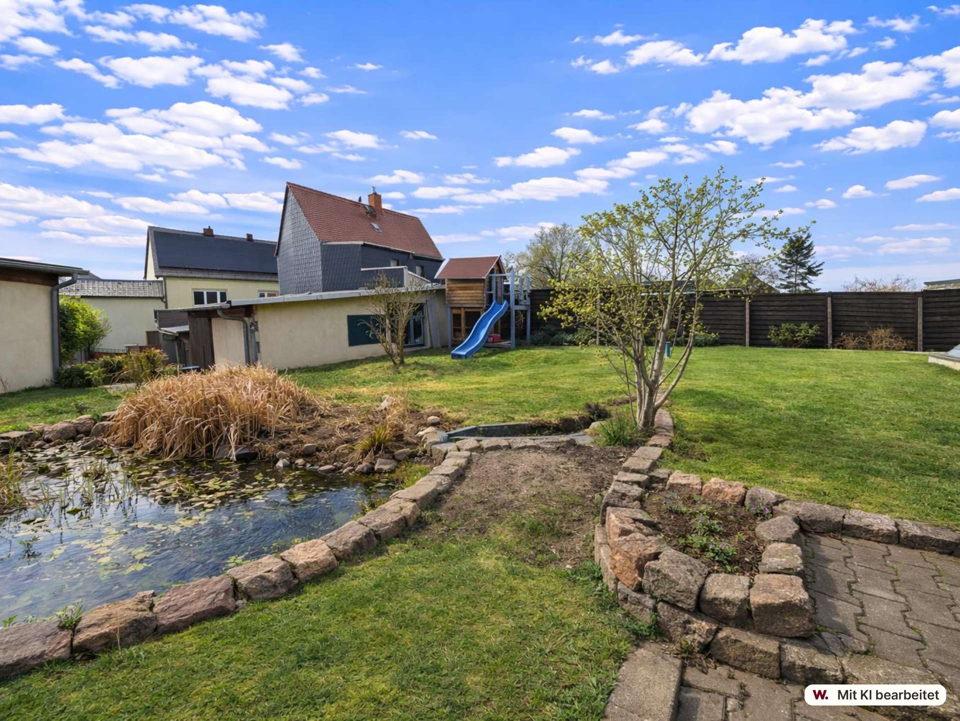 Backyard with a stone-edged pond, a blue playground slide, a small tree, and a wooden fence under a bright blue sky with clouds.
