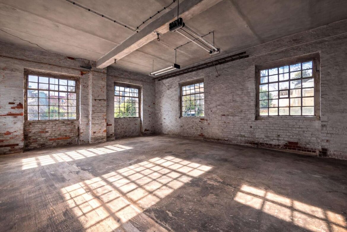 Industrial, empty brick-walled room with large grid windows casting rectangular light patterns on the dusty wooden floor.