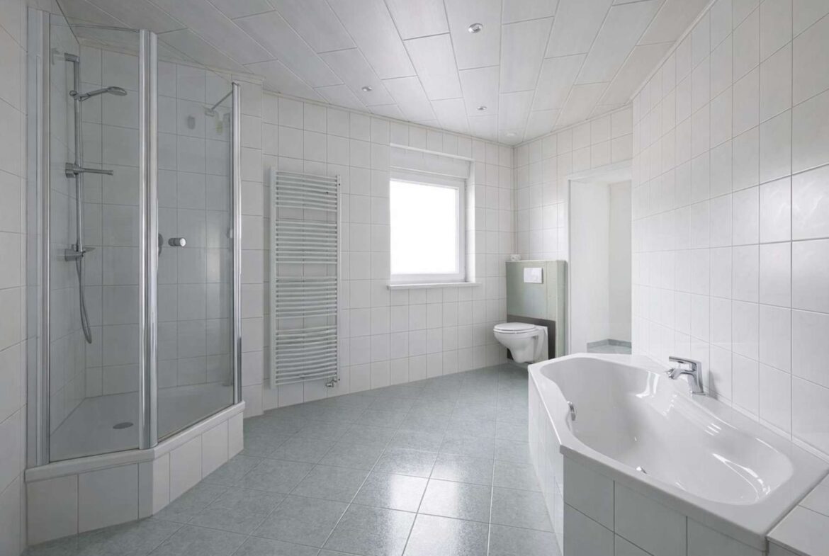 Modern white bathroom with a glass shower enclosure, toilet, and freestanding tub under a bright windowed wall.
