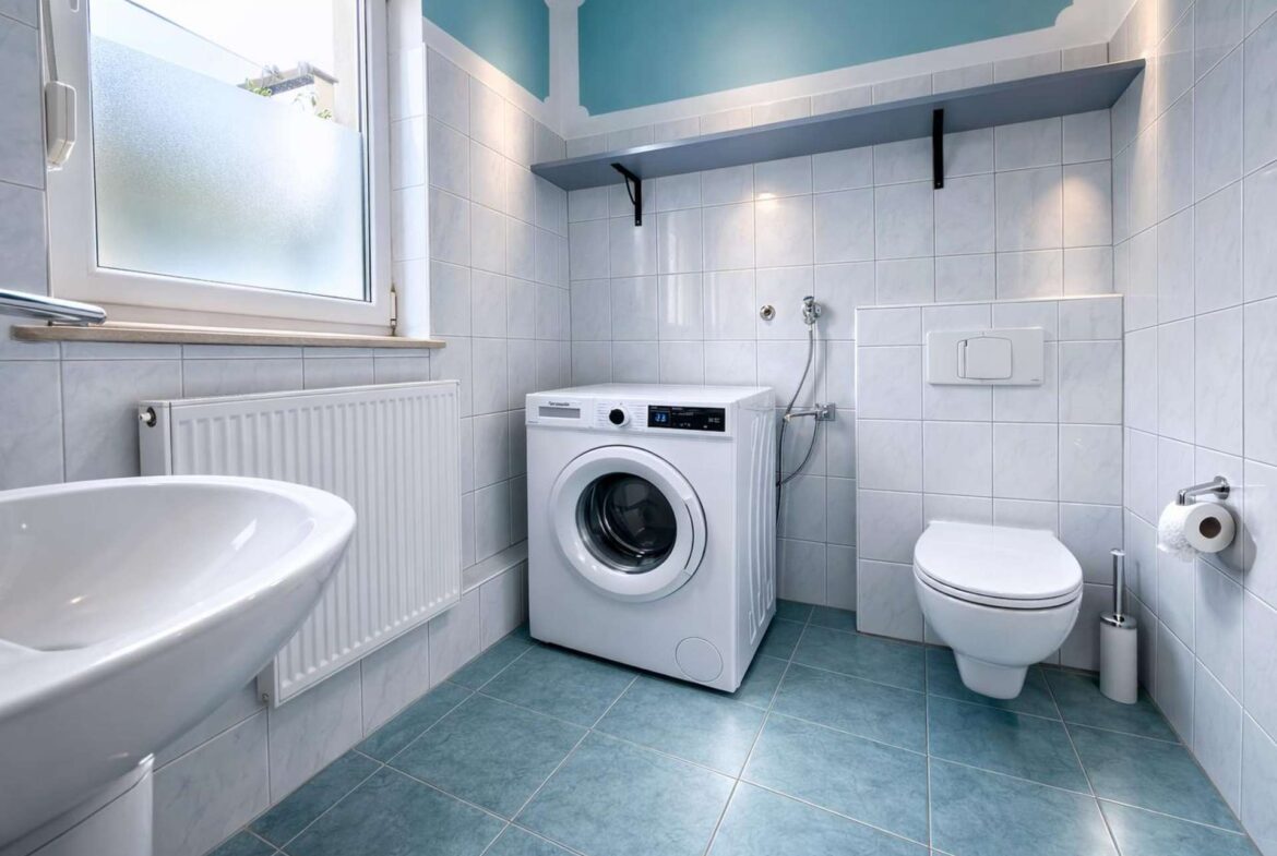 Bathroom featuring a front-loading washing machine next to a wall-hung toilet, sink, and frosted window with blue-tiled walls and floor.