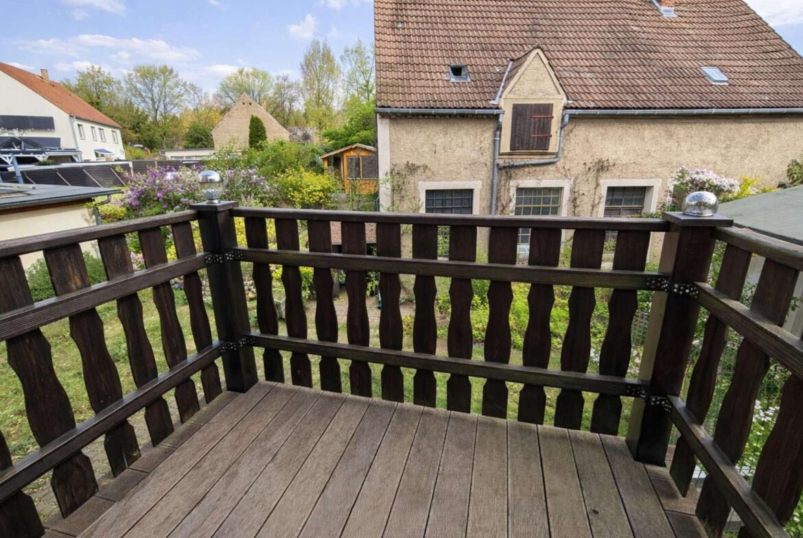 View from a wooden balcony railing toward a beige house with a brown tiled roof and a garden.