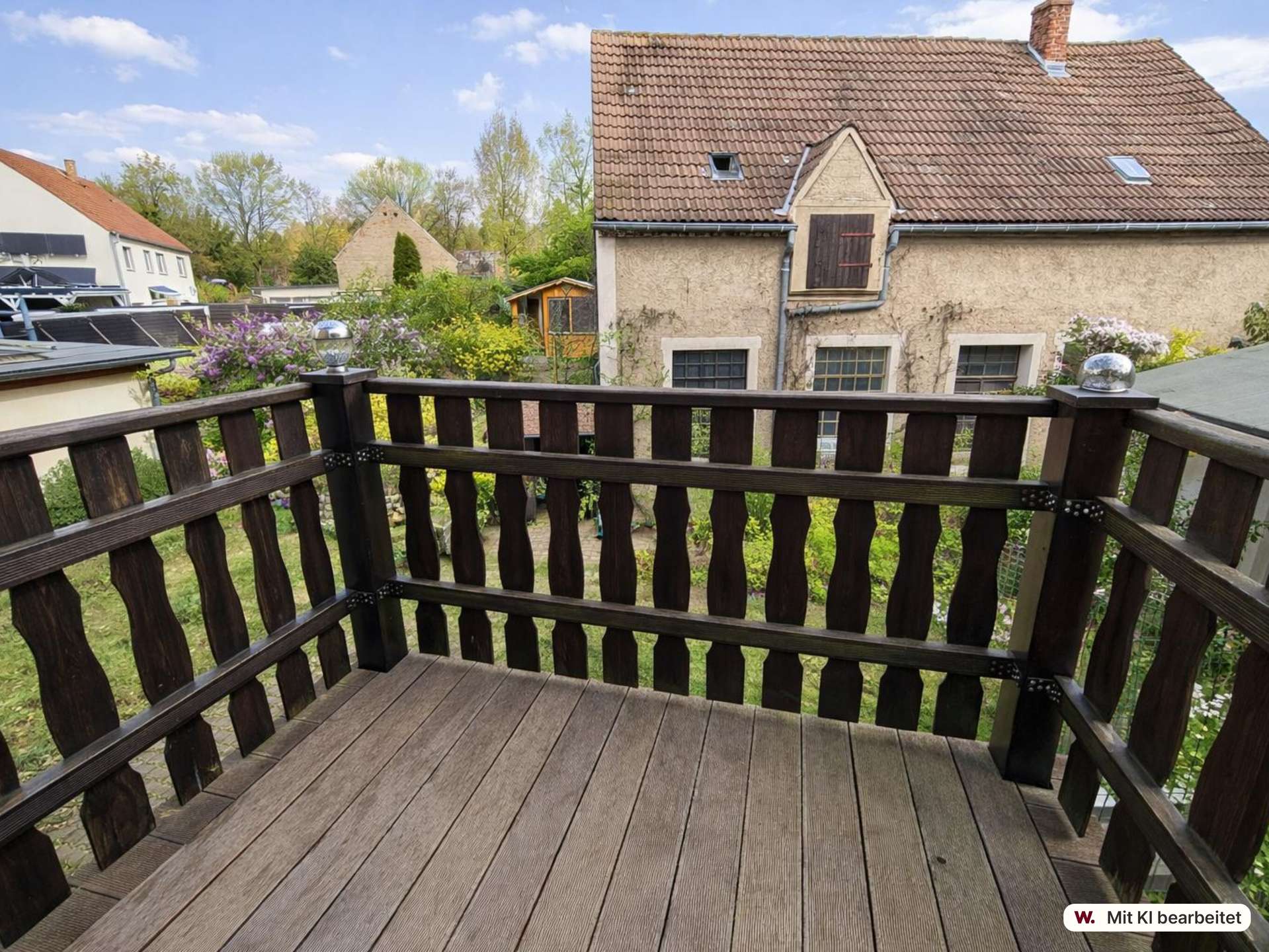 View from a wooden balcony railing toward a beige house with a brown tiled roof and a garden.