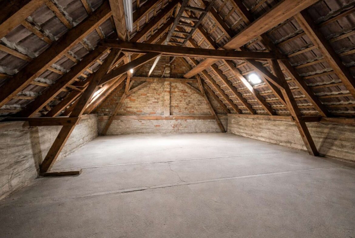 Attic with exposed wooden roof trusses, brick walls, and a bare concrete floor; dim light from small windows.