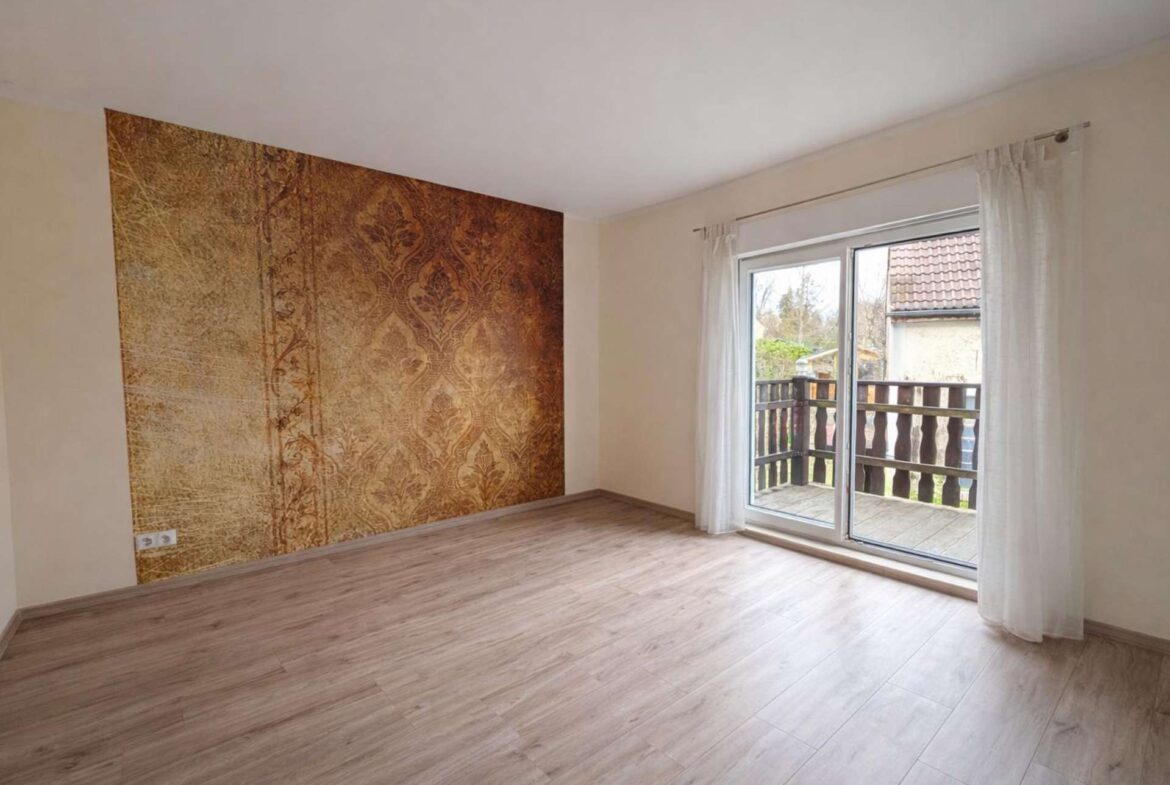 Bright empty living room with a brown ornate wallpaper accent wall, pale walls, light wood flooring, and sliding glass doors opening to a deck with white curtains.