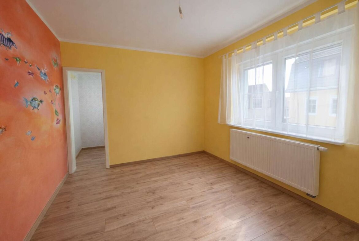 Empty yellow room with an orange accent wall decorated with fish decals, wooden floor, large window with sheer white curtains, and a white radiator beneath the window.