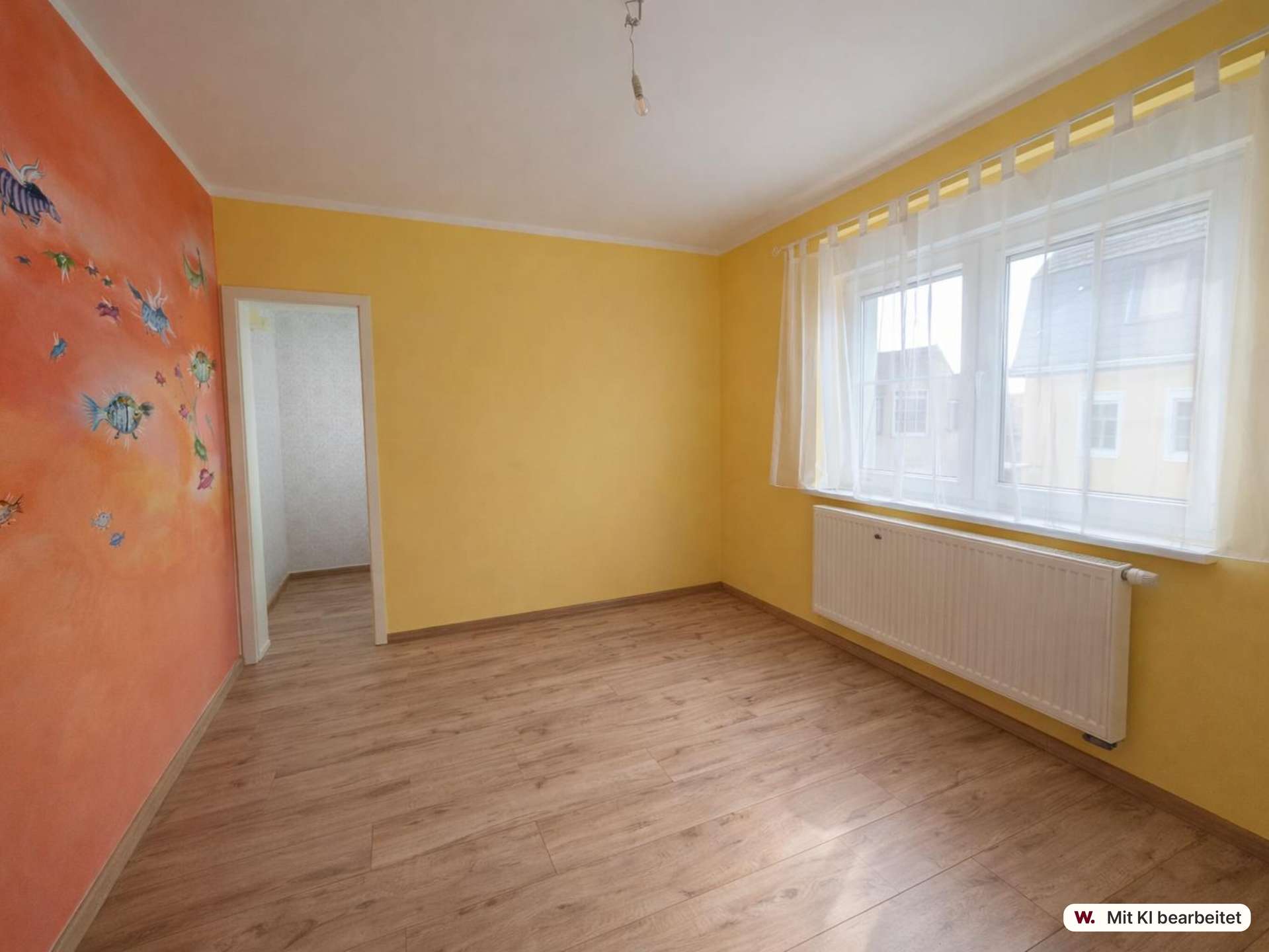 Empty yellow room with an orange accent wall decorated with fish decals, wooden floor, large window with sheer white curtains, and a white radiator beneath the window.