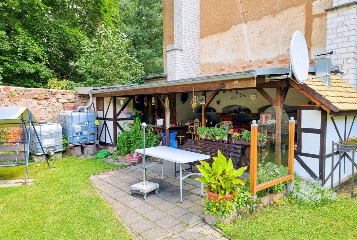 Backyard patio under a wooden awning with a table, bench, chairs, and potted plants alongside a half-timbered wall and satellite dish on the roof.