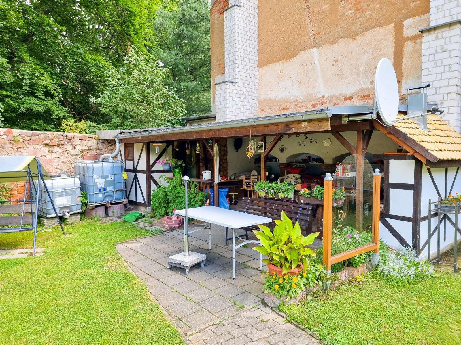 Backyard patio under a wooden awning with a table, bench, chairs, and potted plants alongside a half-timbered wall and satellite dish on the roof.