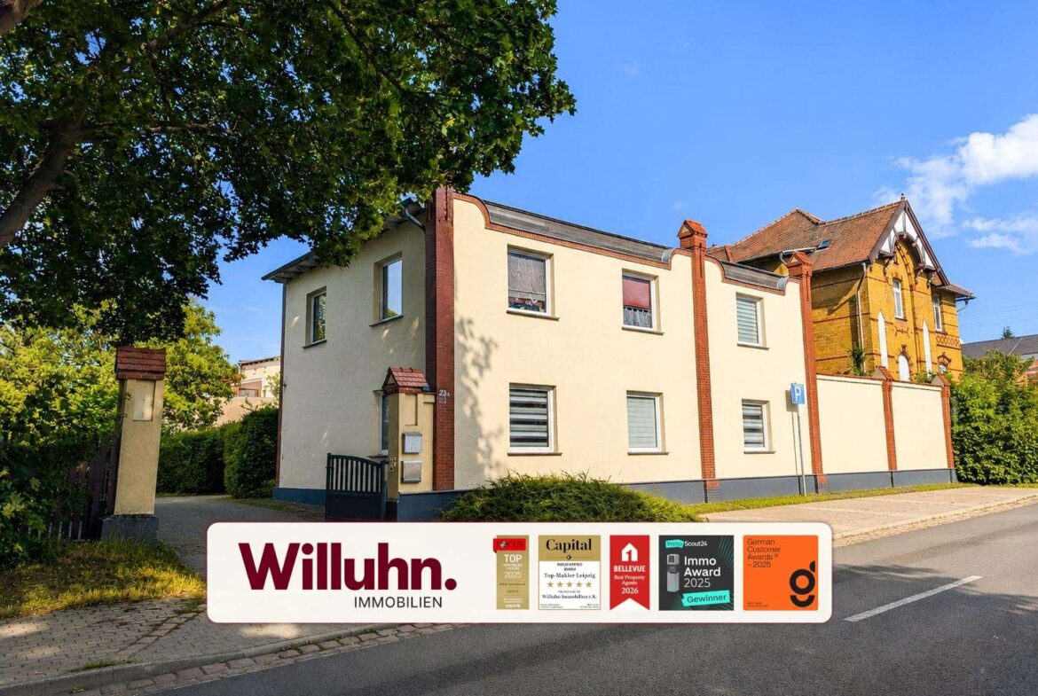 Two-story cream-colored residential building with red brick accents beside a tree-lined street; large promotional banner for Willuhn Immobilien at the bottom edge.