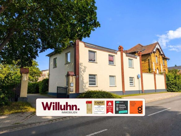 Two-story cream-colored residential building with red brick accents beside a tree-lined street; large promotional banner for Willuhn Immobilien at the bottom edge.