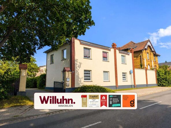 Two-story cream-colored residential building with red brick accents beside a tree-lined street; large promotional banner for Willuhn Immobilien at the bottom edge.