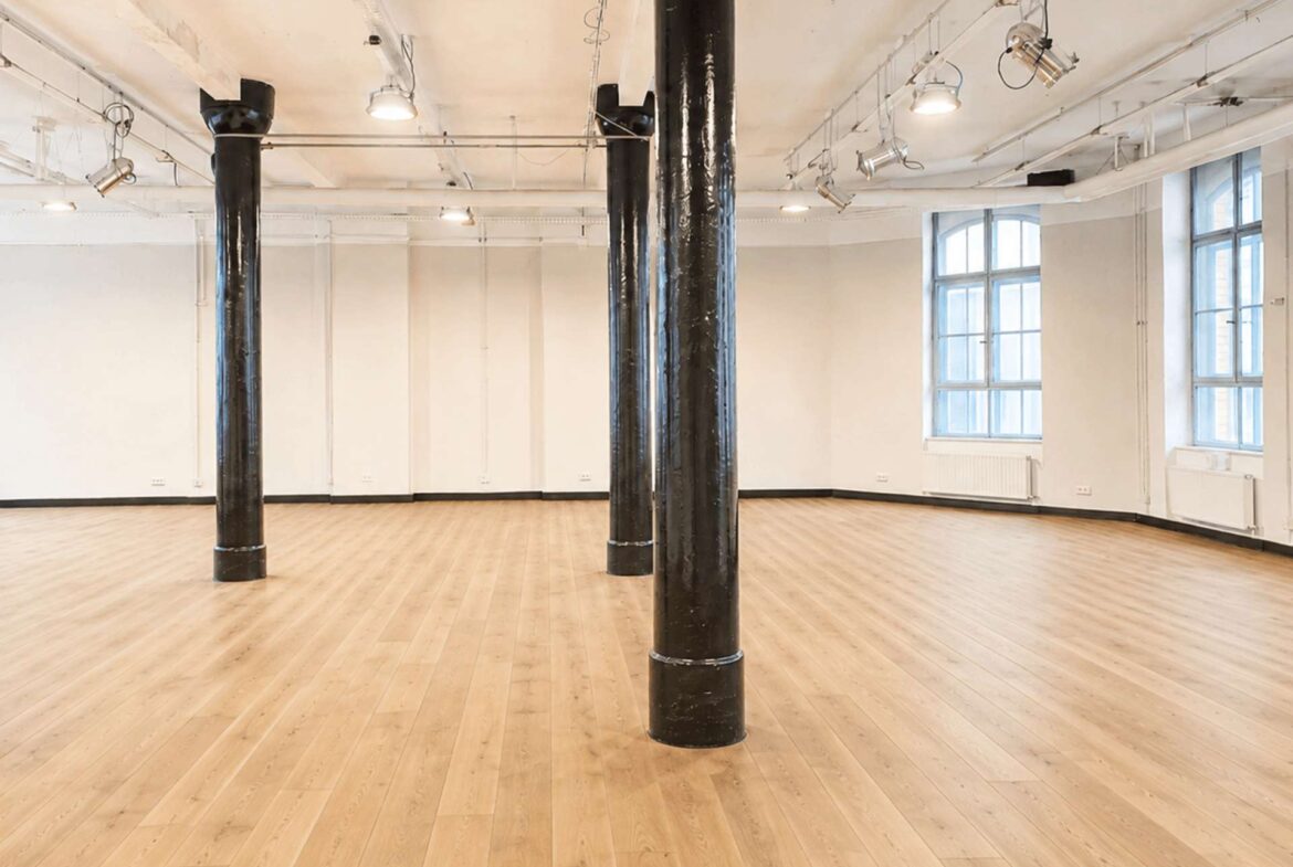 Empty industrial studio with three black pillars, light wood floor, white walls, and large windows; overhead spotlights visible.