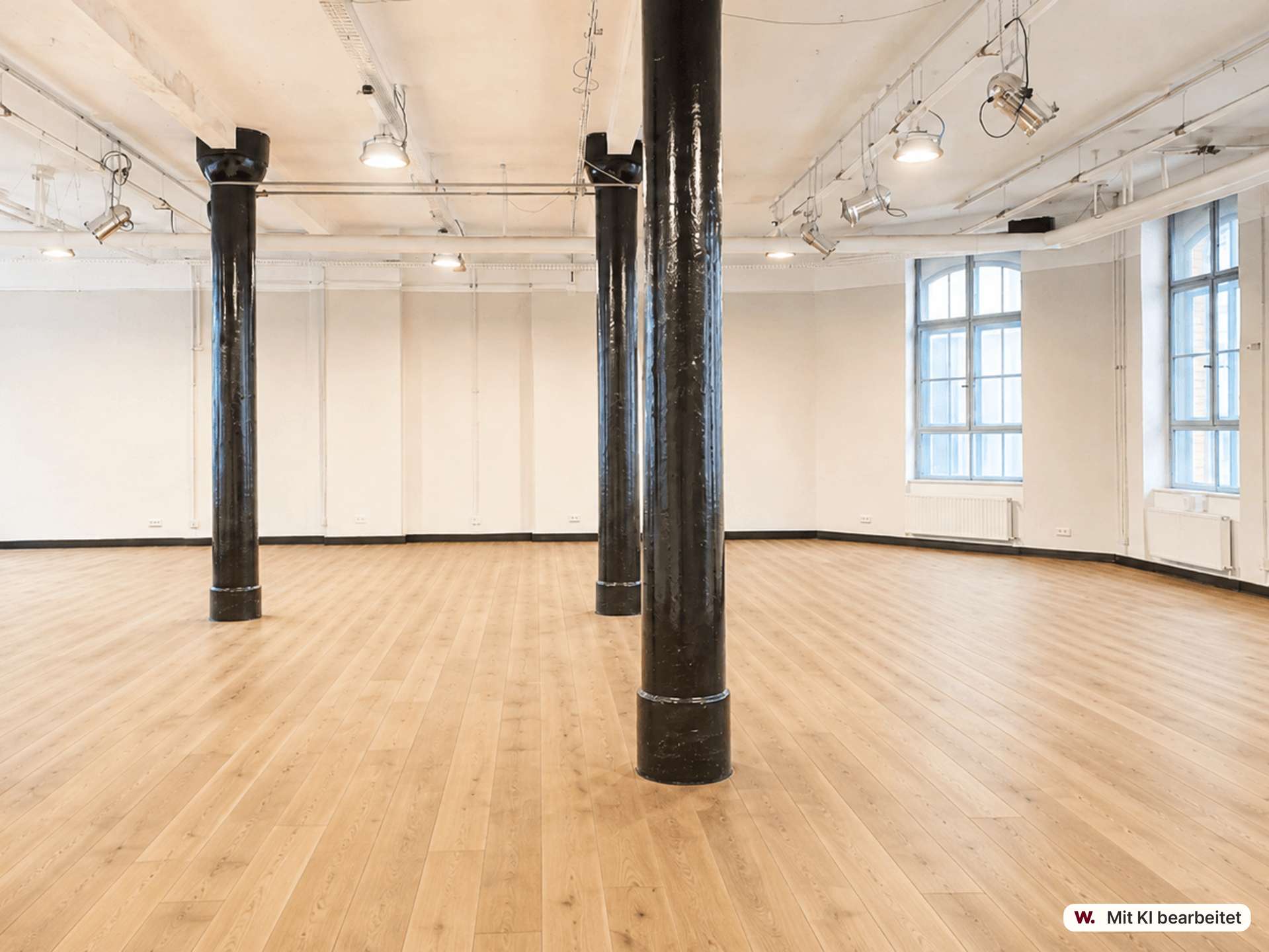 Empty industrial studio with three black pillars, light wood floor, white walls, and large windows; overhead spotlights visible.