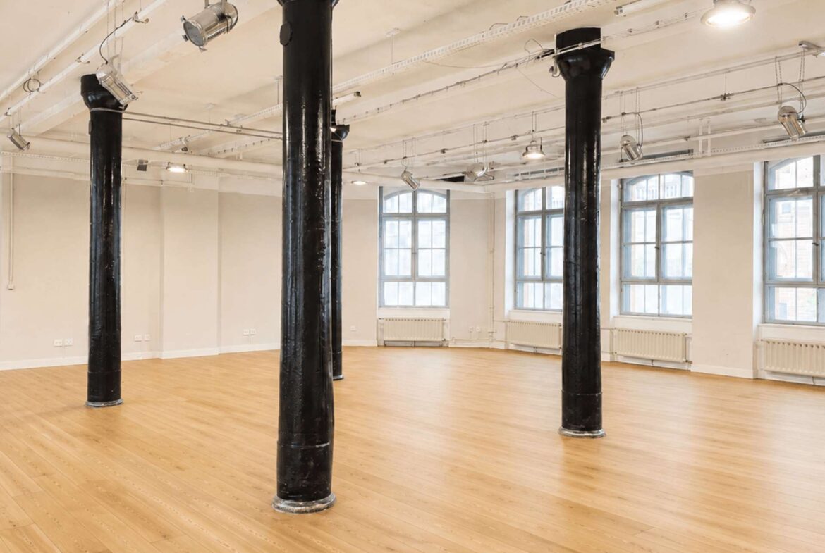 Open studio space with tall black pillars, light wood floor, white walls, and large windows letting in natural light. Overhead track lighting is visible inside the airy room.