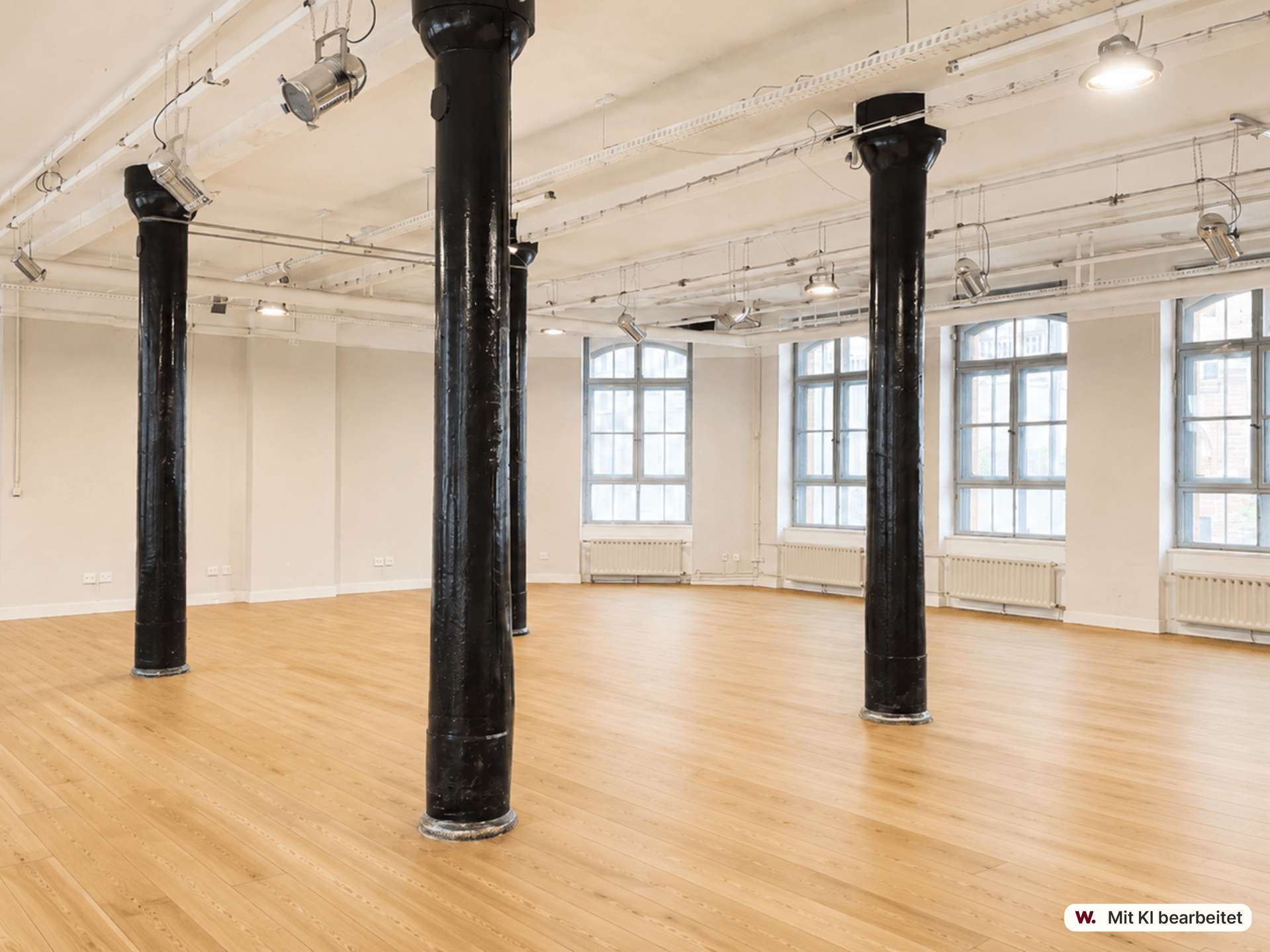 Open studio space with tall black pillars, light wood floor, white walls, and large windows letting in natural light. Overhead track lighting is visible inside the airy room.