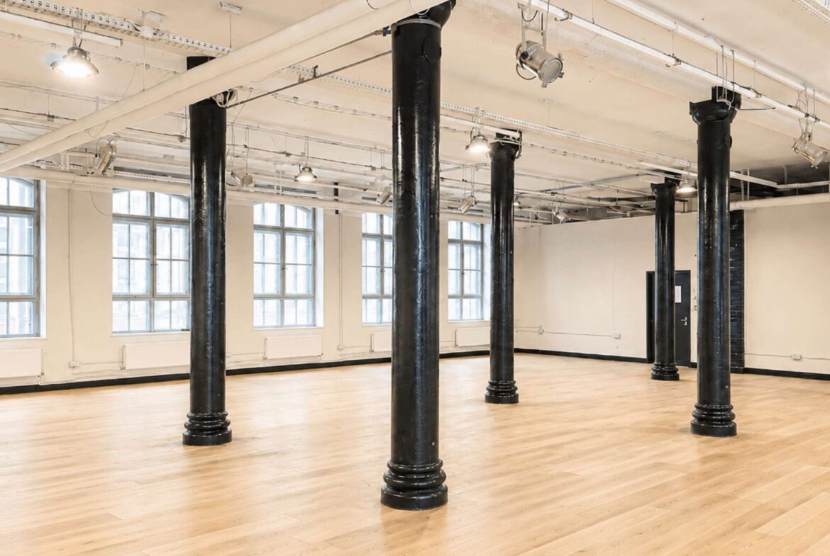 Open industrial studio with black pillars, wooden floor, and large windows letting in natural light.