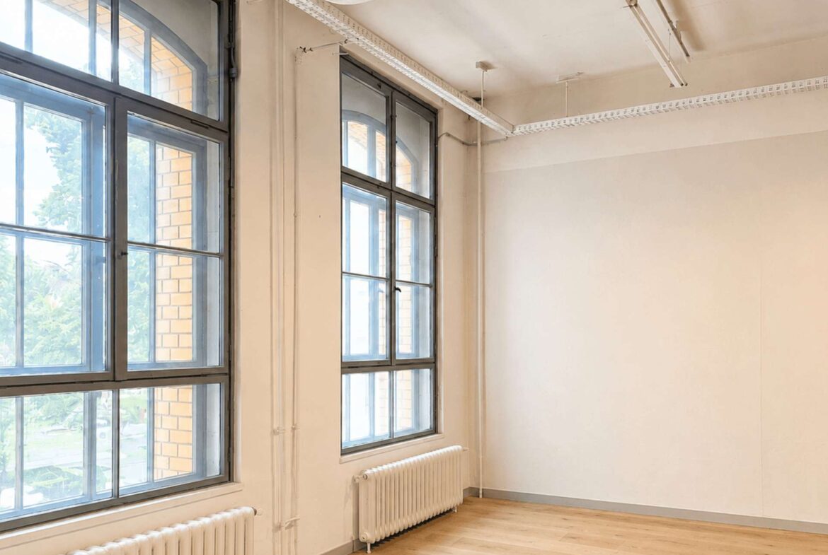 Bright, empty room with tall arched windows and white radiators along the wall, letting in daylight.