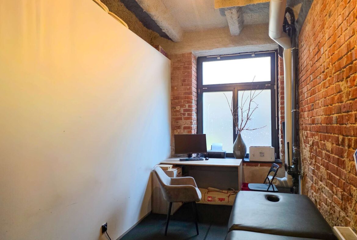 Narrow office corner with brick wall on the right, white wall on the left, and a desk facing a large window by the end.