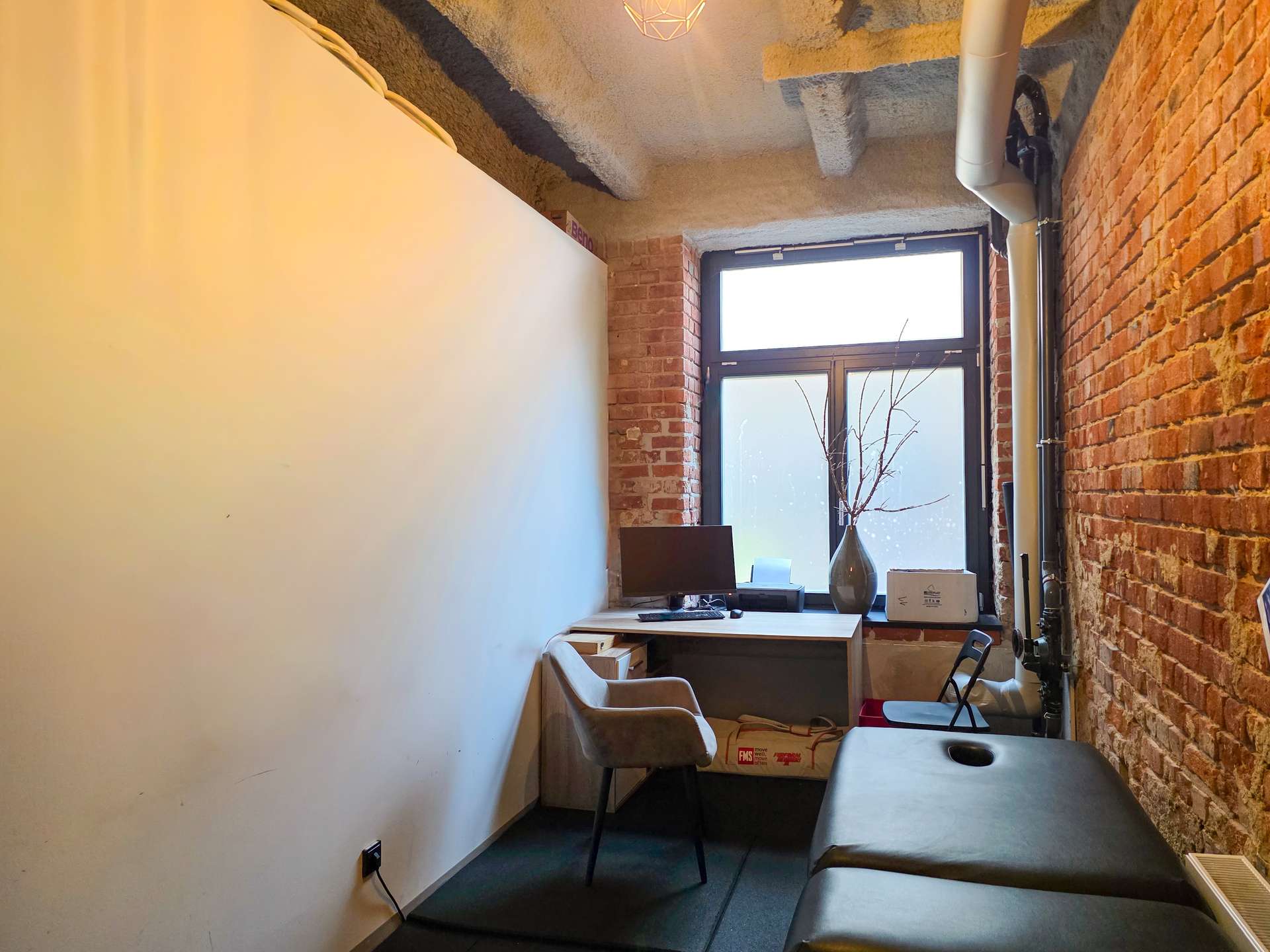 Narrow office corner with brick wall on the right, white wall on the left, and a desk facing a large window by the end.