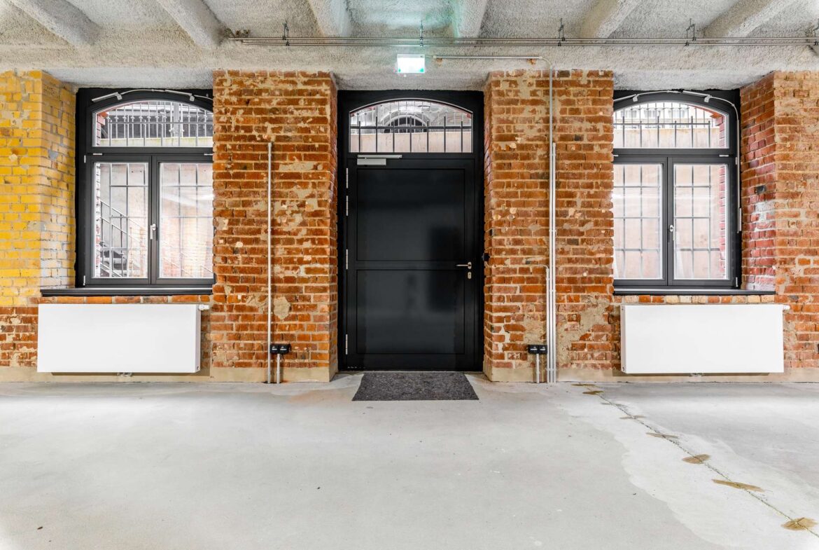 Industrial brick interior featuring a centered black door between two arched, barred windows and exposed pipes above.