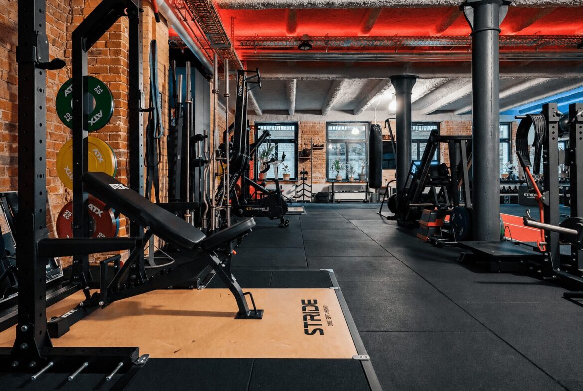 Industrial-style gym interior with weight racks, benches, and colorful plates against brick walls and red overhead lighting.