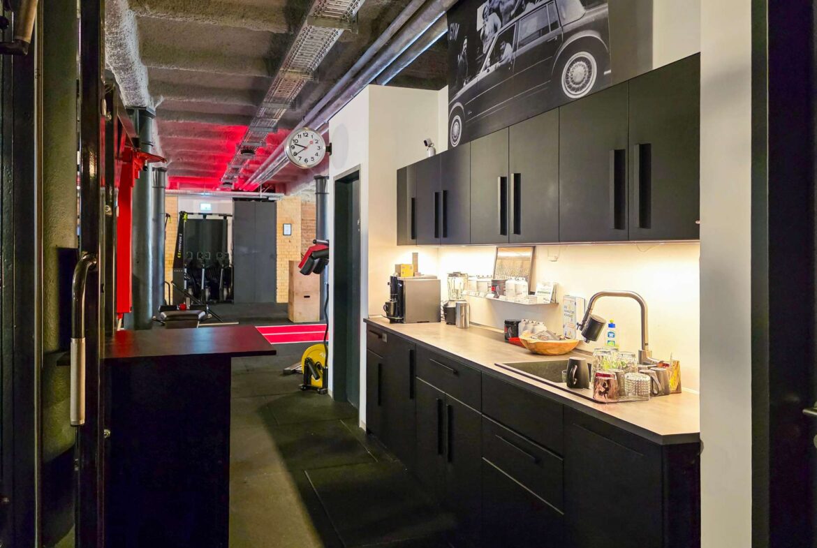 Counter with coffee setup in a modern gym, black cabinets and mugs on the counter, clock on white pillar, and an industrial ceiling with red lighting.