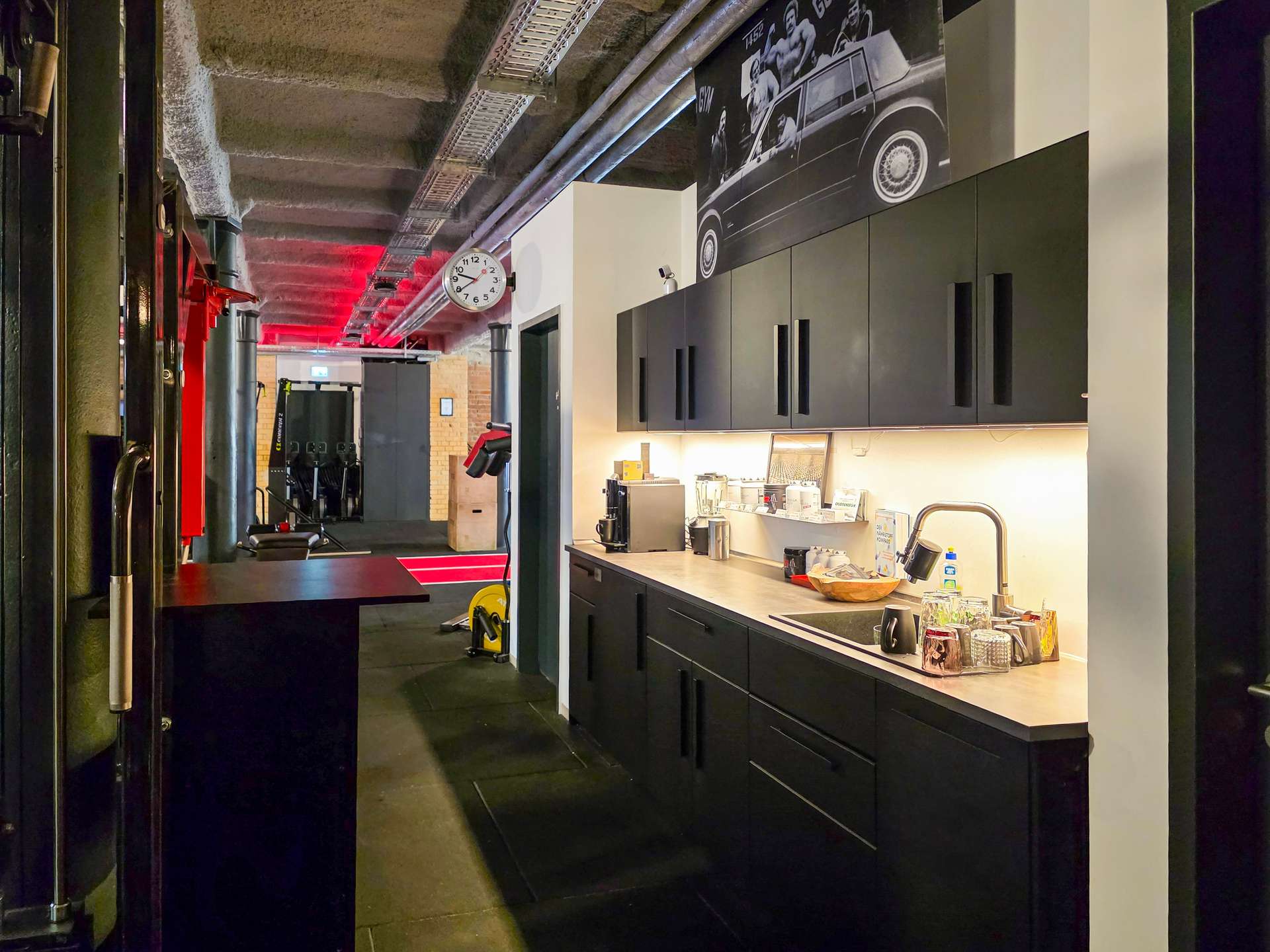 Counter with coffee setup in a modern gym, black cabinets and mugs on the counter, clock on white pillar, and an industrial ceiling with red lighting.