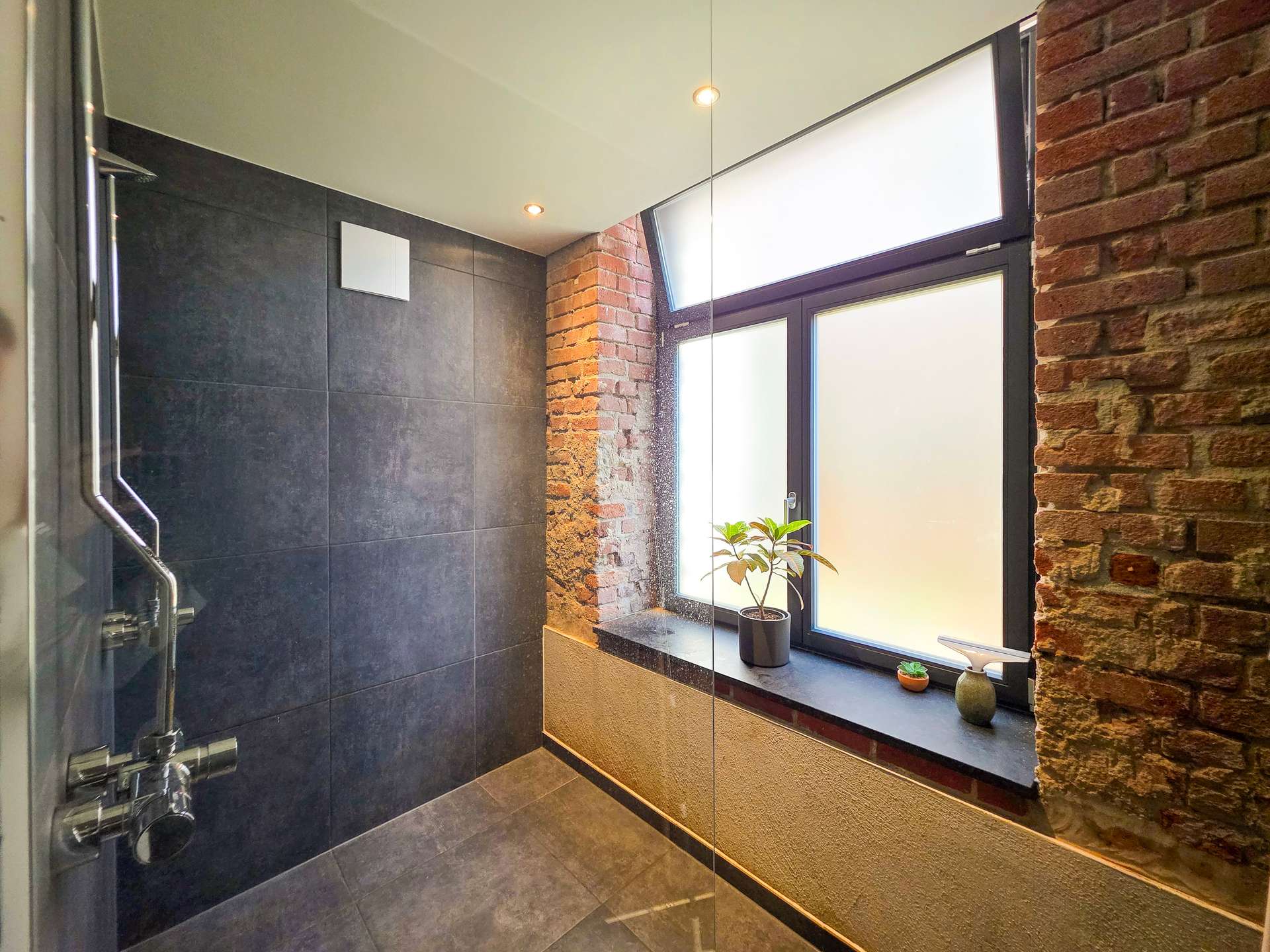 Modern bathroom with dark tiled walls, brick accents, and a glass shower enclosure; frosted window and small plants on the sill.
