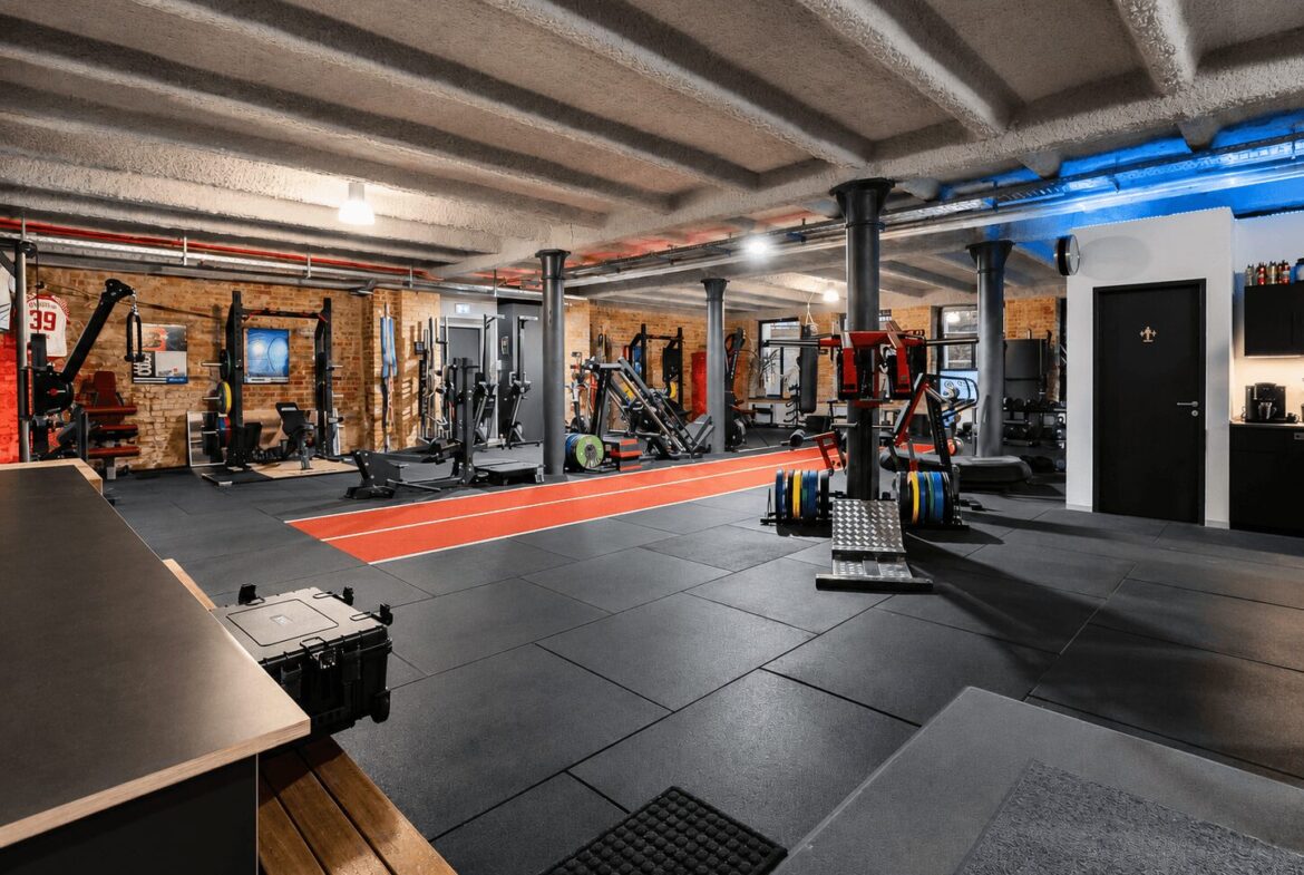 Industrial gym interior with brick walls, exposed ceiling beams, and a red running track surrounded by weight machines.