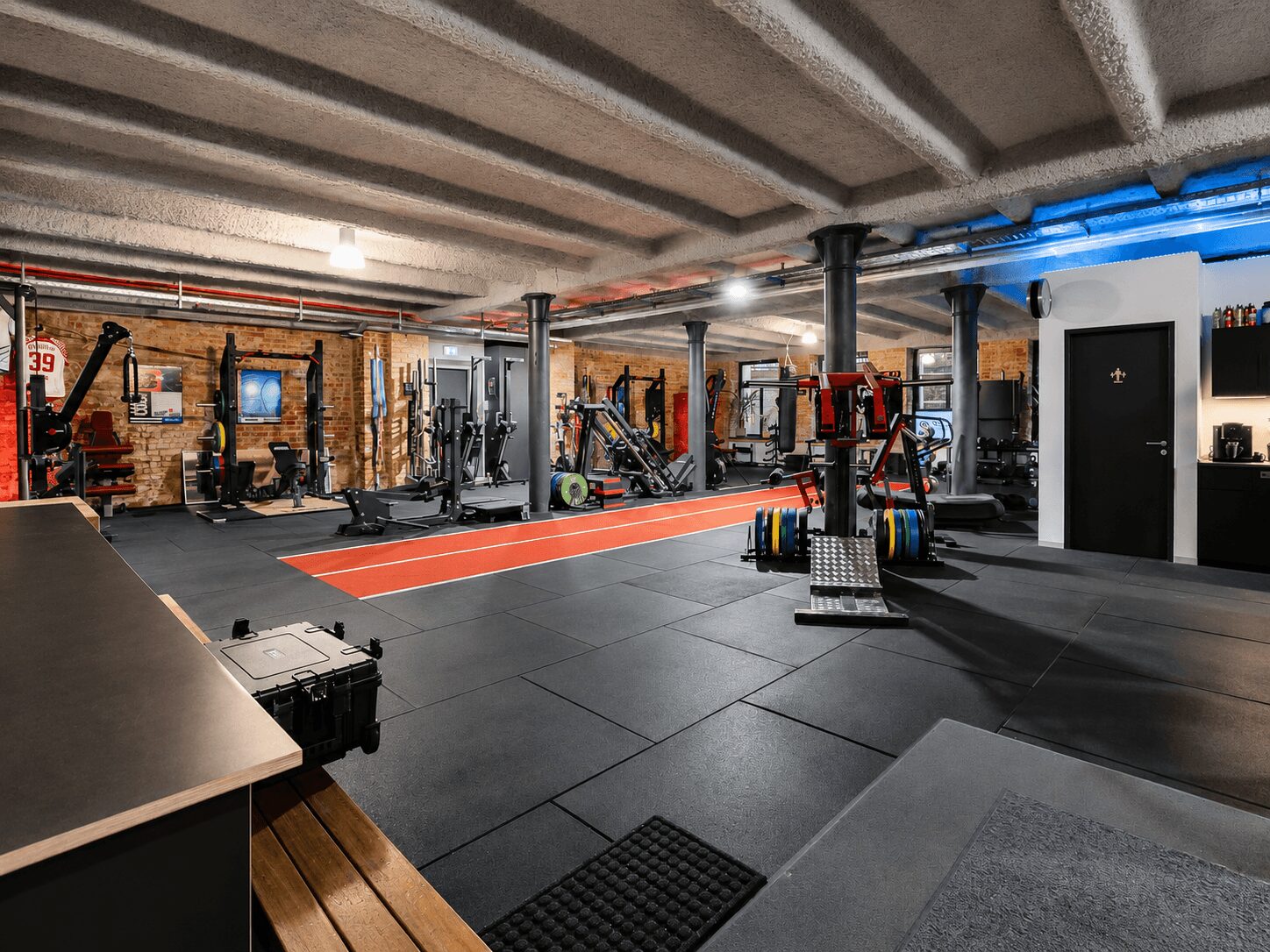 Industrial gym interior with brick walls, exposed ceiling beams, and a red running track surrounded by weight machines.