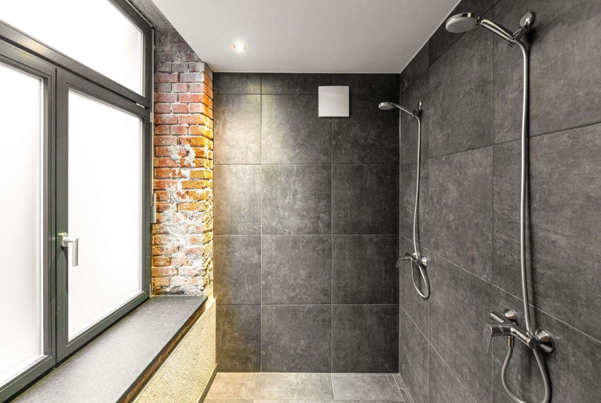 Modern shower area with dark tiled walls, brick accent column, and a large window on the left; dual chrome shower fixtures and handheld sprayer visible.