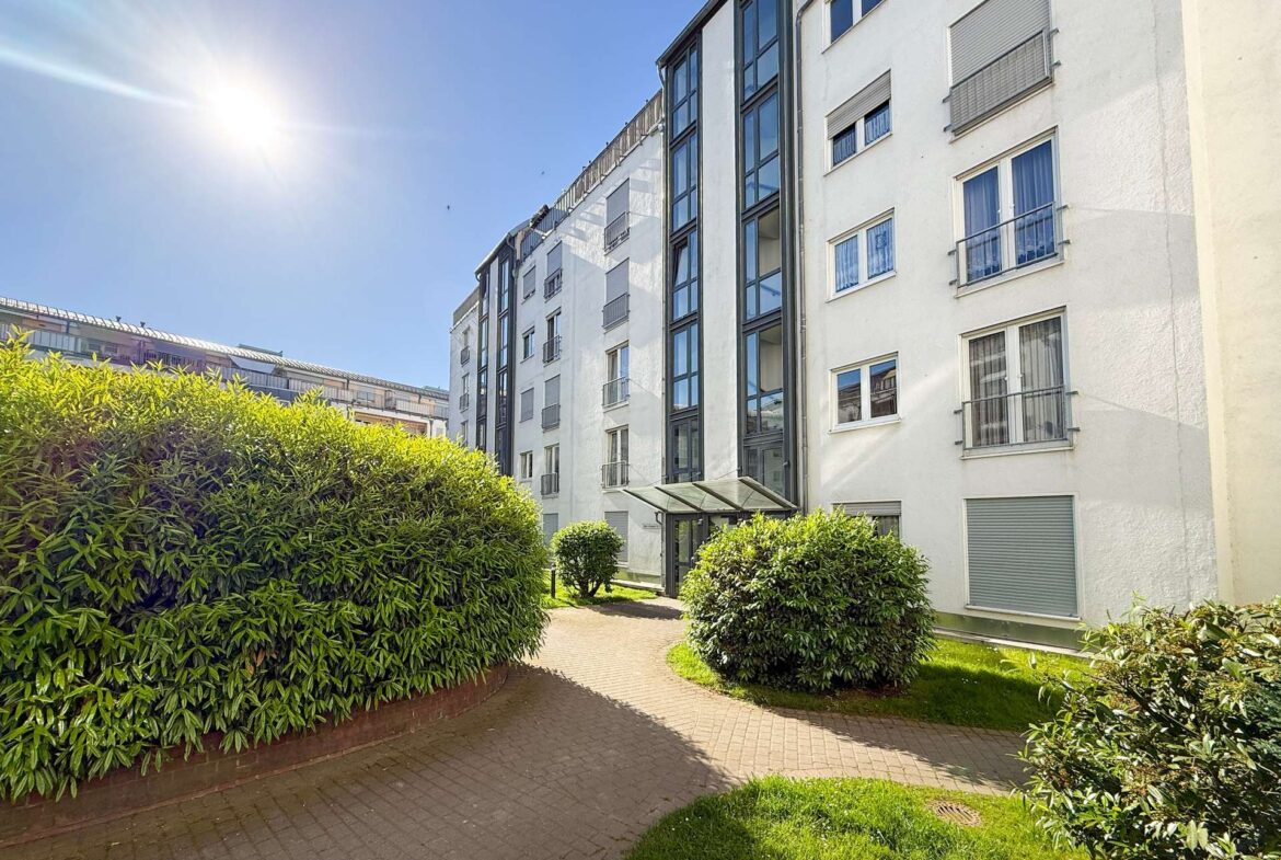 White multi-story apartment building with large glass windows, blue sky, and trimmed hedges along a curved brick path. (Informative image)